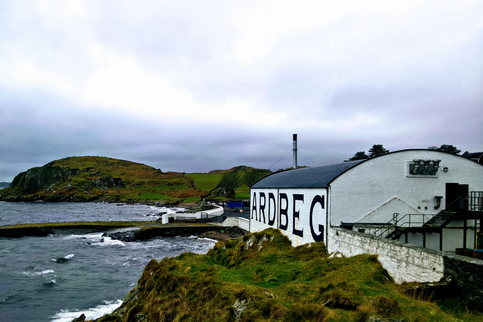 Weiße Ardbeg-Gebäude mit großem Schriftzug unmittelbar an der felsigen Küste.