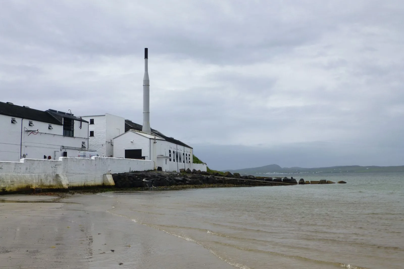 Weiße Gebäude der Bowmore-Brennerei mit Schornstein am Strand des Loch Indaal.