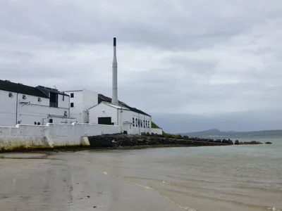 Weiße Gebäude der Bowmore-Brennerei mit Schornstein am Strand des Loch Indaal.